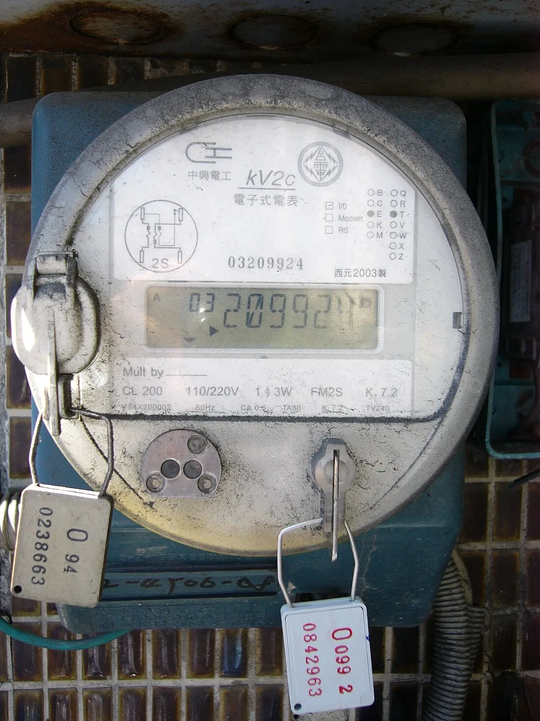 中興電工 kV2c 型電子式電表，面板標示電子式電表、110/220 V 1φ3W、西元 2003 製，液晶顯示器顯示累積度數，表殼下緣掛著兩個編號封印。