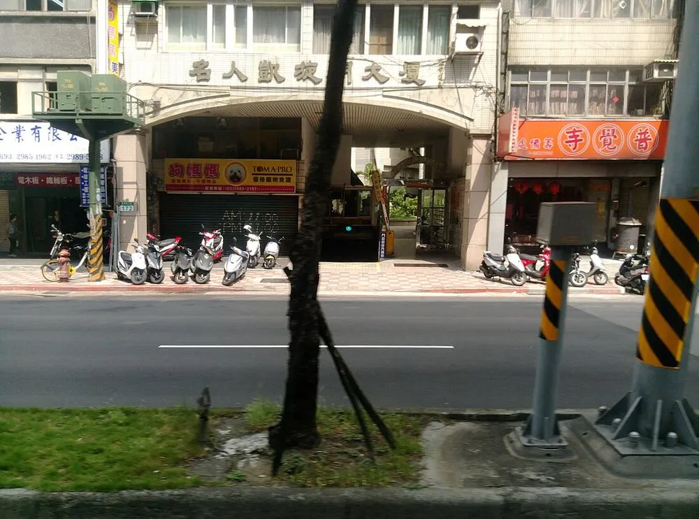 新北市三重區的街景，一棟大樓與騎樓下的店面，路旁有行道樹與電線桿。